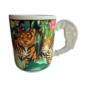 May include: White ceramic mug with a jungle scene featuring two tigers, lush green foliage, and colorful flowers. The handle is sculpted in the shape of an elephant. The mug is a unique piece of drinkware.