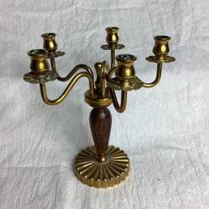 May include: A five-arm candelabra featuring a polished brass finish with a central dark wood stem. The base is circular with a fluted, sunburst-like design, and each of the five candle holders is supported by curved brass arms extending outward.