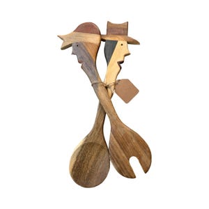 Op de afbeelding: Houten saladeservers met gesneden gezichten, één met een hoed. De servers zijn samengebonden met touw en hebben een blanco label. De lepels zijn gemaakt van licht en donkerbruin hout.