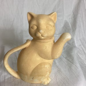 May include: A light beige cat-shaped teapot with a smooth finish. The teapot features a curved handle forming the cat's tail, a spout extending from the cat's raised front paw, and a lid that forms the head of the cat with pointed ears and eyes.