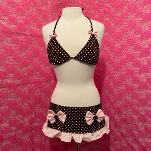 以下が含まれることがあります： マネキンに展示された、茶色と白のドット柄の2ピースビキニセット。ピンクのリボンとフリル付きの裾が特徴です。背景はピンクの花柄です。セットには、ホルターネックのビキニトップとスカートが含まれています。