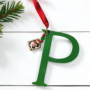 May include: A green wooden letter "P" Christmas ornament with a red ribbon and a small framed photo. The ornament is suspended against a white wooden background, with a sprig of evergreen on the left.