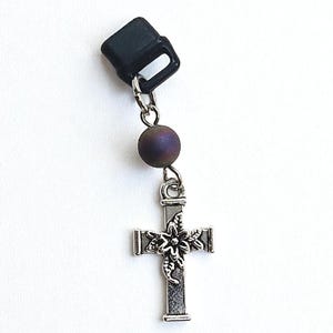 May include: A silver-colored cross pendant with a floral design, connected to a dark purple bead and a black clip. The cross is detailed with intricate patterns and hangs from a small chain.