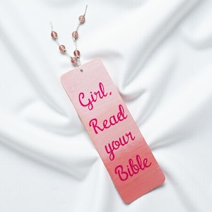 May include: A pink bookmark with the text "Girl. Read your Bible" in a handwritten style. The bookmark has a gradient color scheme, transitioning from light pink to a darker shade. A string of pink beads is attached to the top.