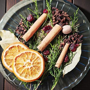 以下が含まれることがあります： 透明なガラスの皿に、乾燥オレンジスライス、シナモンスティック、クローブ、クランベリー、ローリエの葉、緑の小枝をあしらったお祝いの飾り付け。 皿は濃い木製の表面に置かれ、暖かく魅力的な美しさを生み出しています。