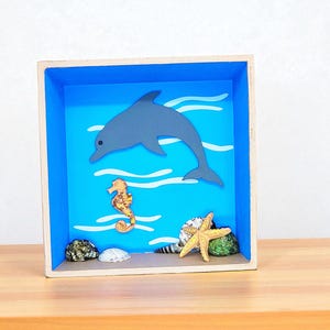 Puede incluir: Una caja de sombra de madera con un fondo azul, que representa una escena marina. Presenta un delfín gris, un caballito de mar y conchas marinas. También se incluye una estrella de mar amarilla y un erizo de mar verde. El marco es de madera.