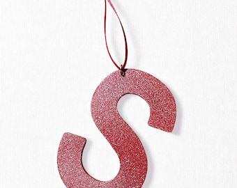 May include: A red glitter-covered letter "S" ornament with a matching red ribbon for hanging. The letter is a bold, stylized design, perfect for holiday or special occasion decor. The ornament hangs against a white background.