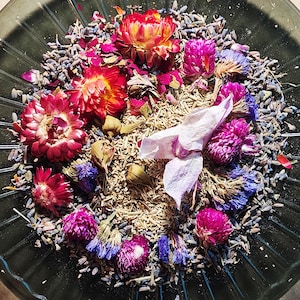 Puede incluir: Primer plano de un cuenco de vidrio lleno de flores y hierbas secas. El arreglo incluye vibrantes flores rojas, rosas y moradas, junto con capullos de lavanda y otros elementos botánicos secos. La composición es una mezcla de texturas y colores.