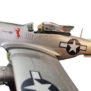 May include: A detailed model of a silver P-51 Mustang fighter plane. The plane features a red horse emblem, a white star within a black circle, and the text "MAJ LEE TAND MILLER". The model is set against a white background.