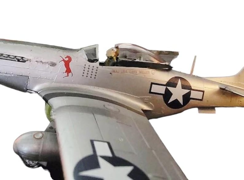 May include: A detailed model of a silver P-51 Mustang fighter plane. The plane features a red horse emblem, a white star within a black circle, and the text "MAJ LEE TAND MILLER". The model is set against a white background.