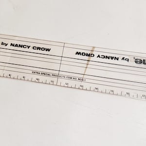 May include: A clear plastic ruler with black markings and the text "Quickline by Nancy Crow." The ruler features measurements in inches, with angled lines for precise cutting. Extra Special Products Item No. MCD-1 is also printed on the ruler.