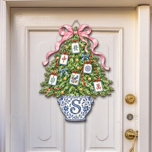 May include: A wall hanging shaped like a green Christmas tree in a blue and white floral pot with the letter S. The tree is decorated with a large pink bow, small gold ornaments, blue ribbons, and several mahjong tiles featuring Chinese characters.