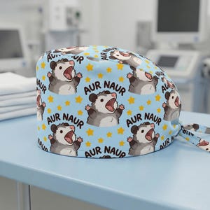 Puede incluir: Un gorro de quirófano azul claro con un patrón repetitivo de zarigüeyas de dibujos animados con la boca abierta y el texto AUR NAUR, intercalado con pequeñas estrellas amarillas. El gorro tiene una corona redondeada y cintas de tela traseras.