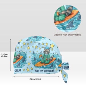 Puede incluir: Un gorro de quir&oacute;fano azul claro con un patr&oacute;n repetitivo de un mapache con uniforme m&eacute;dico sobre una tabla de surf en el oc&eacute;ano, con pizza, estrellas y el texto "AND IT'S NOT GREAT". El gorro mide 13,5 cm de alto y 21 cm de ancho.