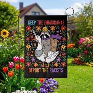 May include: Black garden flag with the text "KEEP THE IMMIGRANTS DEPORT THE RACISTS". The flag features a raccoon wearing a crown and cape riding a goose, surrounded by colorful flowers.