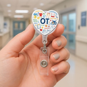 以下が含まれることがあります： 青い文字「OT」が描かれた、心臓型のバッジリール。作業療法に関連するカラフルなアイコンとテキストで囲まれています。リールは透明なプラスチックストラップと格納式のコードを備えています。