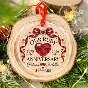 May include: A round, clear glass ornament with a wood-grain background, featuring a red heart graphic, a red ribbon bow, and the text "OUR RUBY ANNIVERSARY 1975 2026 Patricia Isabella 51 YEARS". It hangs from a red ribbon loop.