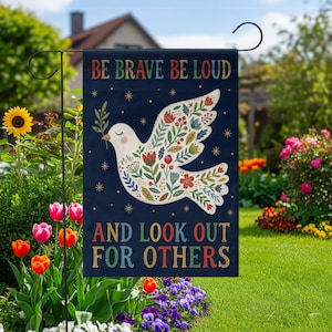 Könnte beinhalten: Eine Gartenflagge mit marineblauem Hintergrund zeigt eine weiße Taube mit floralen Designs und dem Text "BE BRAVE BE LOUD AND LOOK OUT FOR OTHERS" in bunten Buchstaben. Die Flagge wird in einem Garten gezeigt.