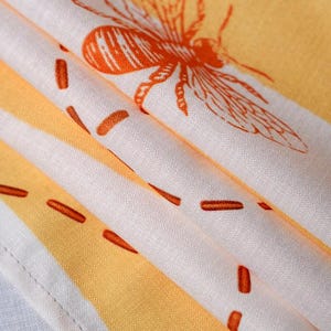 May include: A folded textile featuring a repeating pattern of orange bees and dashed lines printed on a white and yellow striped background. The fabric has a visible woven texture and a stitched hem along the bottom edge of the material.