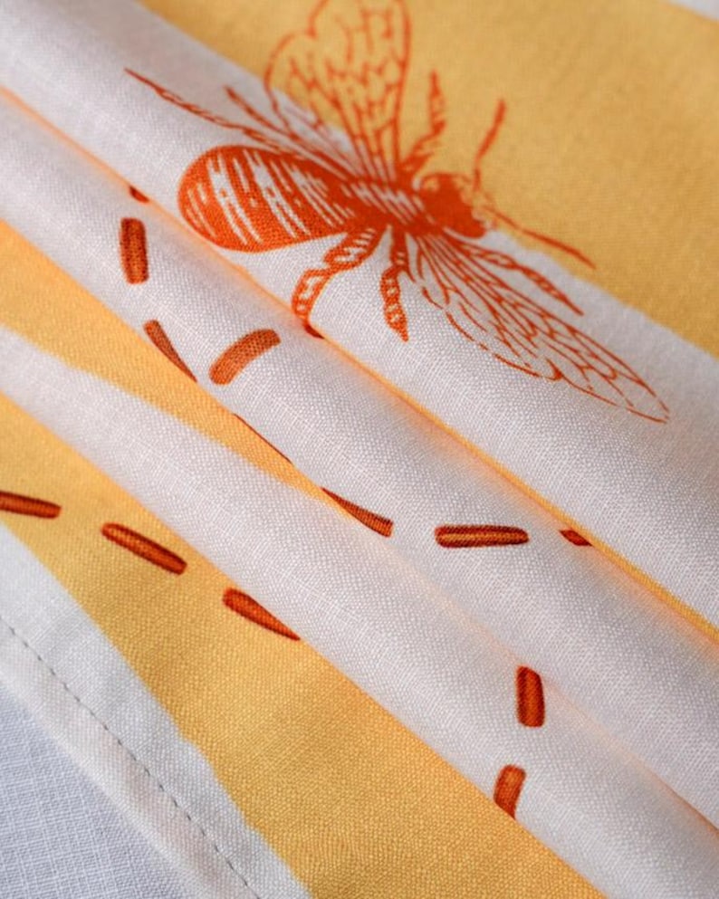 May include: A folded textile featuring a repeating pattern of orange bees and dashed lines printed on a white and yellow striped background. The fabric has a visible woven texture and a stitched hem along the bottom edge of the material.