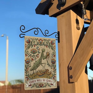 May include: A decorative garden flag featuring a detailed illustration of two praying mantises with floral accents. The flag has the text "GIRLS WILL BE GIRLS" on a banner. The flag is hanging from a black metal bracket.