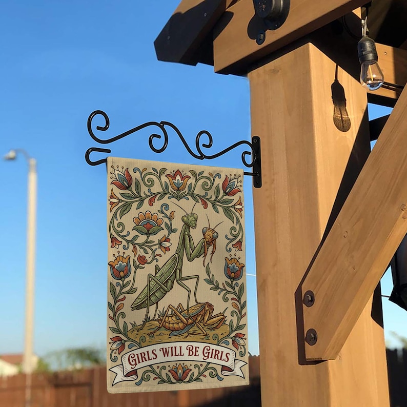 May include: A decorative garden flag featuring a detailed illustration of two praying mantises with floral accents. The flag has the text "GIRLS WILL BE GIRLS" on a banner. The flag is hanging from a black metal bracket.