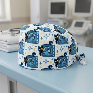 May include: A white surgical cap with a repeating pattern of black cat silhouettes, cat skeleton x-rays, and the text "CAT SCAN" in blue. The design also includes blue and yellow stars. The cap is tied at the back.