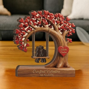 May include: A wooden Valentine's Day decoration featuring a tree with red heart-shaped leaves and a swing with two silhouettes. A heart with the names "Kristin Andrea" is on the trunk. The base reads "Our first Valentine 02.10.2026".