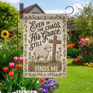 May include: A beige garden flag with the text "Even in the Chaos, His Grace Still Finds Me." The flag features a lamb, a cross, and floral accents. The flag is displayed on a black metal garden stake in a garden setting.