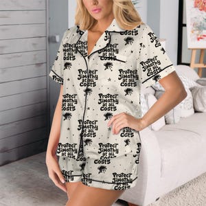 May include: A cream-colored short-sleeve pajama set with black piping, featuring a repeating pattern of small black star icons and the text "Protect Jimothy at all costs" printed in a bold, black, sans-serif font across the shirt and shorts.