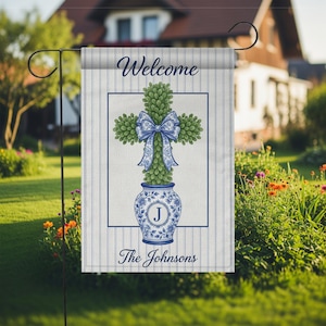 Könnte beinhalten: Eine Gartenflagge mit blau-weiß gestreiftem Hintergrund. Sie zeigt einen grünen, kreuzförmigen Formschnitt mit einer blauen Schleife, platziert in einer blau-weißen Vase. Die Wörter "Welcome" und "The Johnsons" sind aufgedruckt.