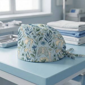 May include: A light blue surgical scrub cap featuring a botanical and skeletal design. The pattern includes illustrations of bones, flowers, and leaves in shades of blue, green, and cream. The cap is tied at the back.
