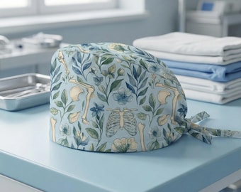 May include: A light blue surgical scrub cap with a botanical and skeletal design. The pattern includes illustrations of bones, flowers, and leaves in shades of blue, green, and cream. The cap is tied at the back.