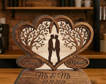 May include: A dark wood decorative stand featuring two interlocking heart-shaped frames with tree branch cutouts. Inside the hearts are silhouettes of two people holding hands. The base is engraved with "Mr & Mr" and "02.10.2026", with "Tommy" and "Michael" on side plaques.