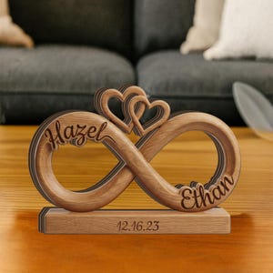 May include: A wooden infinity symbol ornament featuring two interlocking hearts at the top. The name Hazel is engraved on the left loop, Ethan on the right loop, and the date 12.16.23 is engraved on the rectangular wooden base stand.