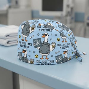 May include: Light blue scrub cap with a repeating pattern of a goose wearing sunglasses and a vest, holding a coffee cup and an X-ray. The text "IDK, JUST TAKE A PICTURE" is also printed on the cap. The cap has a tie in the back.