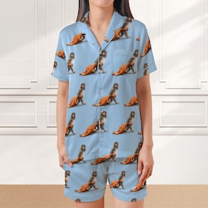 May include: Light blue pajama set featuring a repeating pattern of a monkey carrying a fried chicken leg. The set includes a short-sleeved button-up shirt and matching shorts. The fabric has a silky sheen.