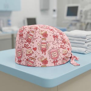 May include: A pink surgical cap with a repeating pattern of medical illustrations, including IV bags, medicine bottles, hearts, and roses. The cap features the text "Levo Epi" and "Atropine".