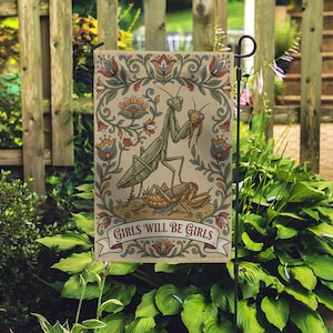May include: A garden flag featuring a detailed illustration of two praying mantises and a grasshopper, set against a beige background with floral accents. The flag includes the text "GIRLS WILL BE GIRLS" on a banner.