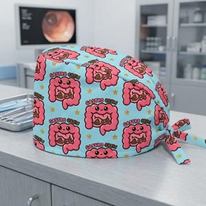 May include: A light blue surgical scrub cap featuring a repeating pattern of smiling pink intestines with brown poop and yellow stars. The words "Gastro Crew" are printed on the cap. The cap is tied at the back with a matching ribbon.