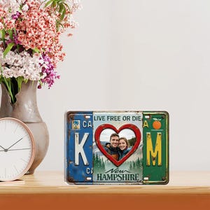 May include: A license plate-style photo frame with a heart-shaped cutout. The frame is divided into sections with the letters K and M, and a heart-shaped photo of a couple. The text "LIVE FREE OR DIE" is visible.