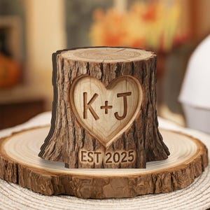 Könnte beinhalten: Holzbaumstumpf-Dekoration mit einem geschnitzten Herz mit den Initialen "K+J" und dem Text "EST 2025". Der Stumpf ist braun mit einem strukturierten Rindenmuster und steht auf einem Holzsockel.