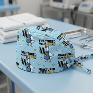 May include: A light blue surgical cap with a repeating pattern of cartoon geese wearing sunglasses and holding X-ray images. The text "FRACTURED MEANS BROKEN" is also printed on the cap, along with star shapes.