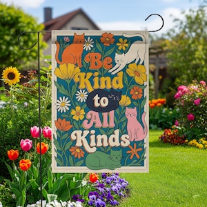 Puede incluir: Una bandera de jardín con el texto "Be Kind to All Kinds" en un estilo retro, rodeada de flores coloridas y gatos en naranja, blanco, rosa, negro y verde. La bandera se muestra en un soporte de jardín de metal negro.