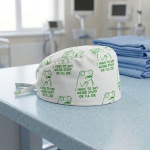 May include: White surgical scrub cap with a repeating pattern of green cartoon frogs and the text "I HAVE TO SAY WEIRD STUFF OR I'LL DIE." The cap is tied at the back and sits on a light blue surface.