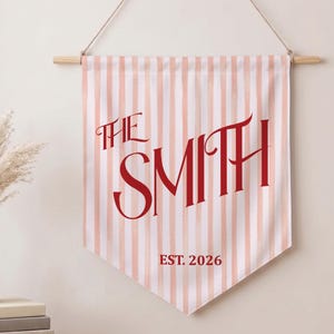 May include: A fabric wall banner featuring vertical light pink and white stripes. The text "THE SMITH" is printed in large, bold red lettering across the center, with "EST. 2026" printed in smaller red text below. The banner hangs from a wooden rod.
