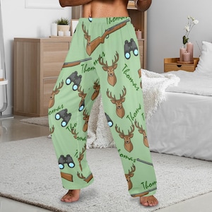 May include: Light green pajama pants with a repeating pattern of deer heads, rifles, and binoculars. The name "Thomas" is written in a script font throughout the design. The pants have a relaxed fit and a comfortable appearance.