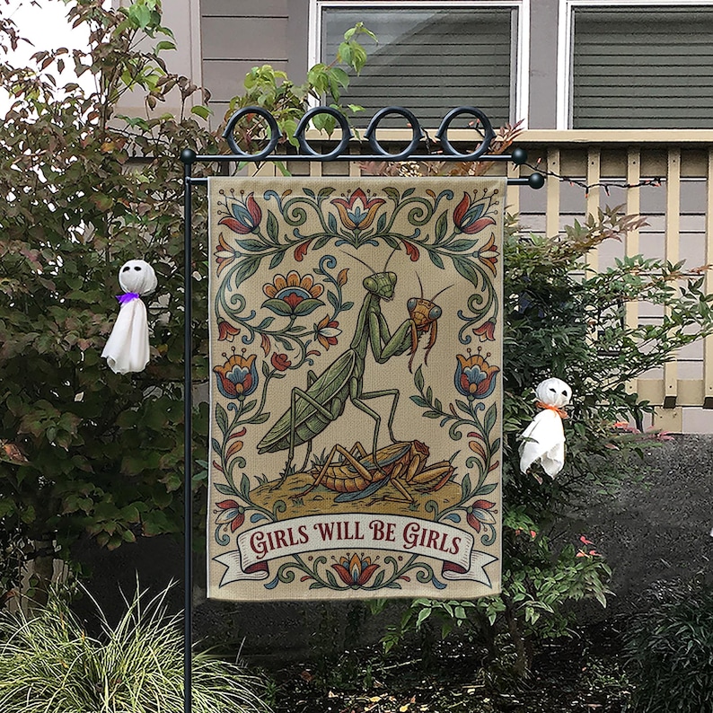 May include: A beige garden flag featuring a detailed illustration of a praying mantis with a floral border. The flag reads "GIRLS WILL BE GIRLS" in a banner. The flag is displayed on a black metal stand, with two small ghost decorations.