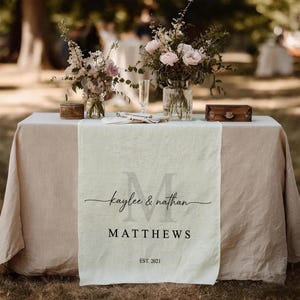 May include: A table setting with a cream-colored table runner featuring the names "Kaylee & Nathan" and the last name "MATTHEWS" with "EST. 2021". The table is set with floral arrangements and a small wooden box.