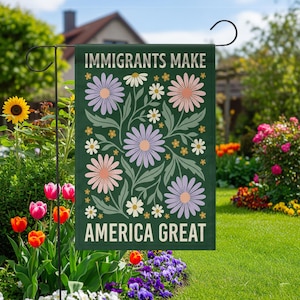 以下が含まれることがあります： 「IMMIGRANTS MAKE AMERICA GREAT」の白い文字が入った濃い緑色のガーデンフラッグ。紫とピンクの大きな花、白いデイジー、緑の葉のフローラルデザインが特徴です。庭に飾られた旗です。